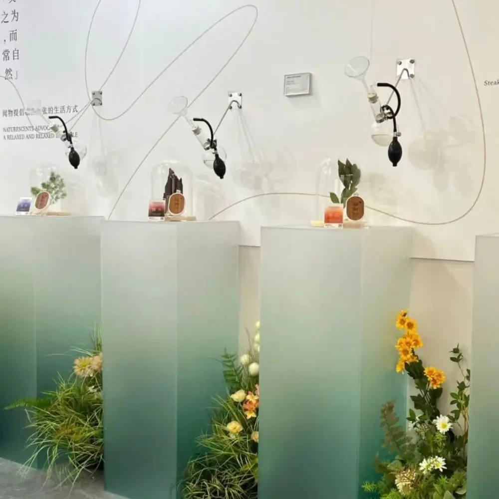 Gradient frosted acrylic display plinths featuring a green ombre finish, supporting glass cloches and botanical samples in an interactive scent discovery station with wall-mounted manual diffusers.